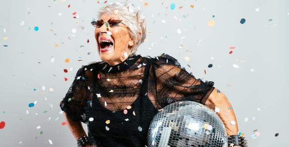 Mevrouw met grijs haar en een zonnebril op. Ze heeft een discobal in haar hand en lacht heel breed. Er is confetti te zien